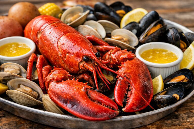Close-up landscape image of a classic Maine seafood platter featuring a bright red cooked lobster in the foreground, surrounded by steamed clams and mussels, with small bowls of melted butter and lemon wedges on a rustic wooden table.